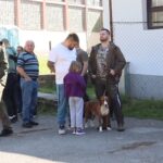 Skup lovaca i posjetilaca na prvoj izložbi lovačkih pasa 'Istinijada 2026' u Bronzanom Majdanu, sa vidljivim psima na povocu i školom u pozadini.
