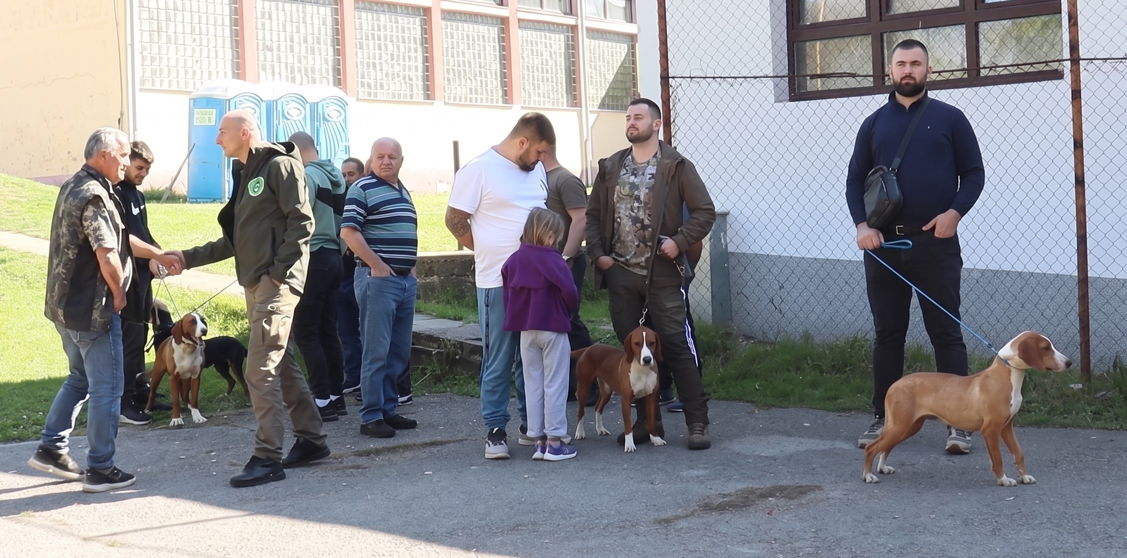 Skup lovaca i posjetilaca na prvoj izložbi lovačkih pasa 'Istinijada 2026' u Bronzanom Majdanu, sa vidljivim psima na povocu i školom u pozadini.